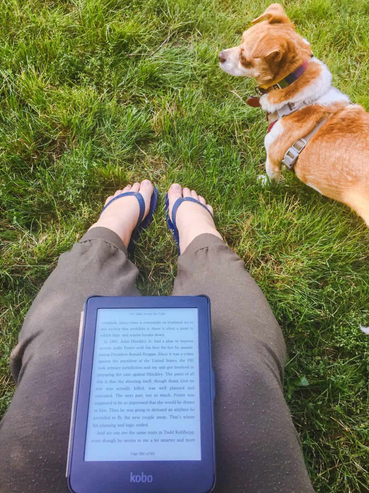 40+ Beach Reads for Every Reading Mood 30 Shot of Riana's legs with an ebook in her lap and her dog Ellie on the grass in front of her