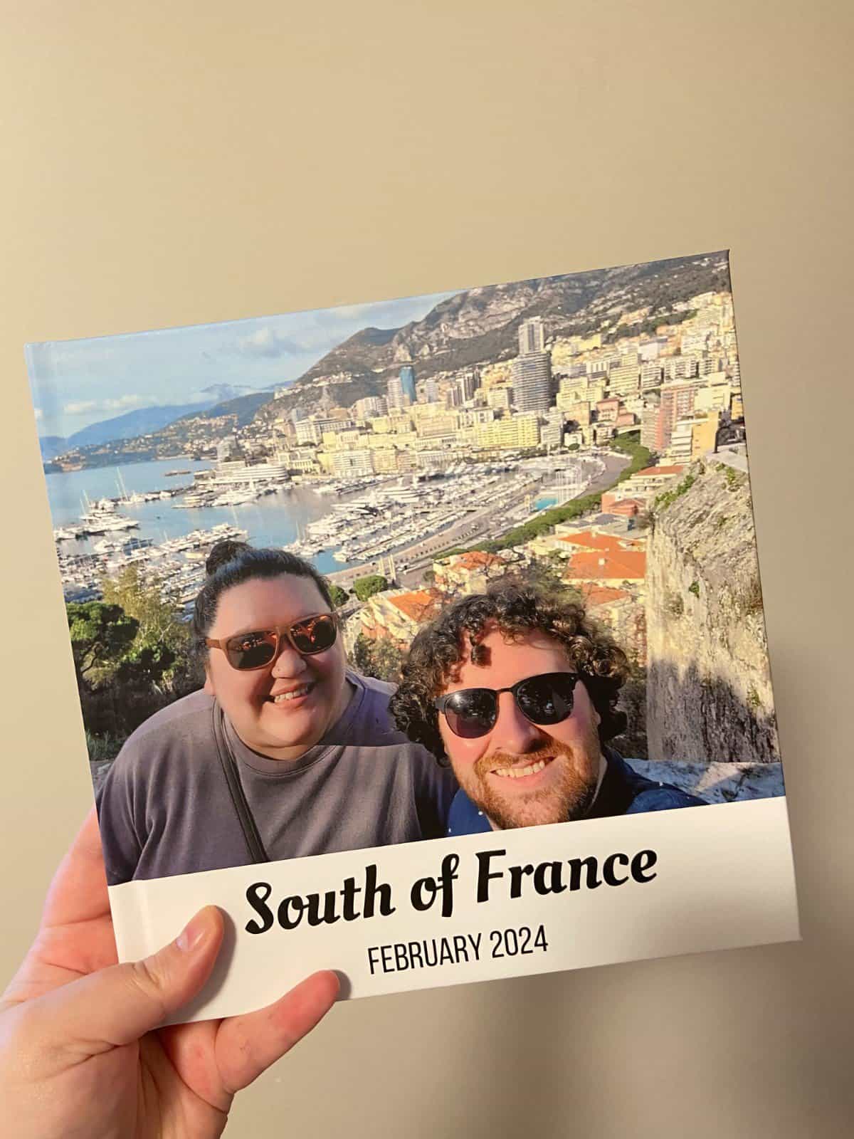 Riana's hand holding a photo book called South of France February 2024 featuring a selfie of Riana and Colin in Monaco; Mixbook photo books review post