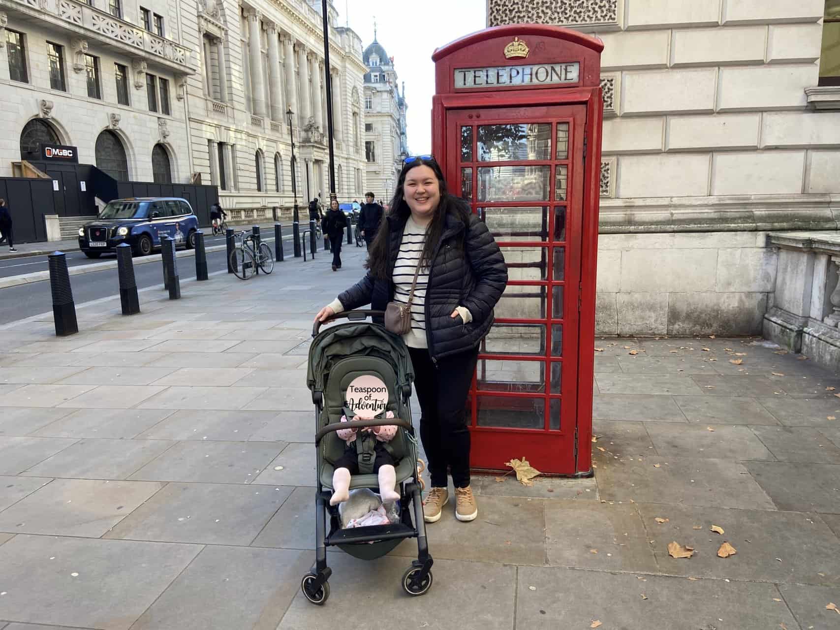 Riana, owner and writer at Teaspoon of Adventure travel blog, standing next to a red phone booth in London with her daughter, Baby C in a stroller in front of her