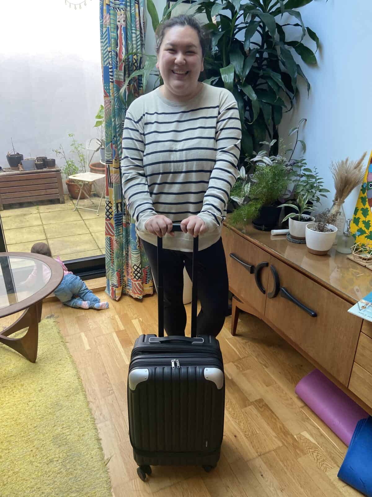 Our Essential Packing List for Travel with a Baby 10 Riana posing with the LEVEL8 Grace 20" carry-on suitcase while Baby C crawls in the background