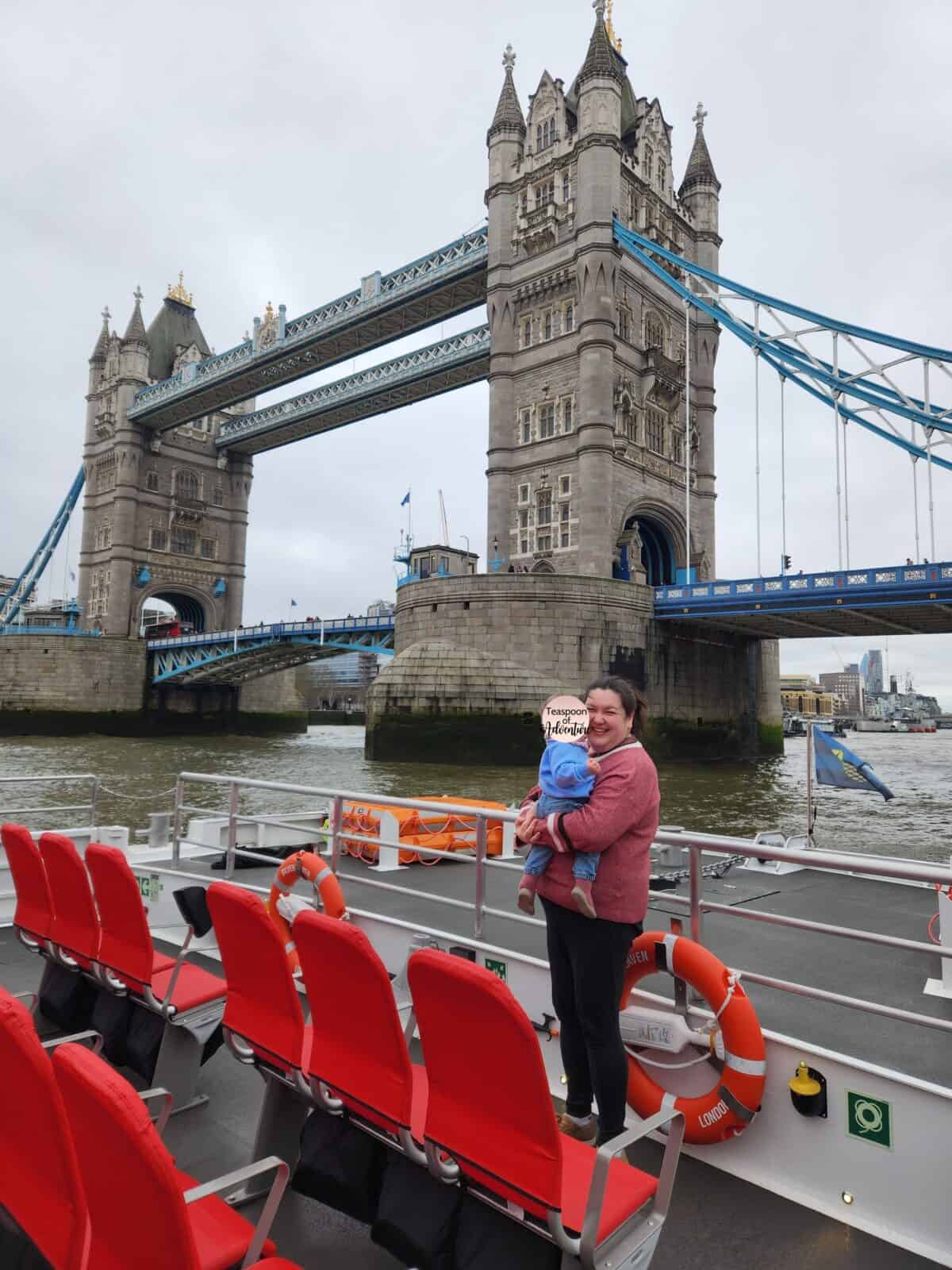 20+ Things to Do in London With a Baby 36 Riana holding Baby C at the bow of the SIlver Raven electric boat with the Tower of London River Tour Thames River cruise. The Tower of London is behind them.