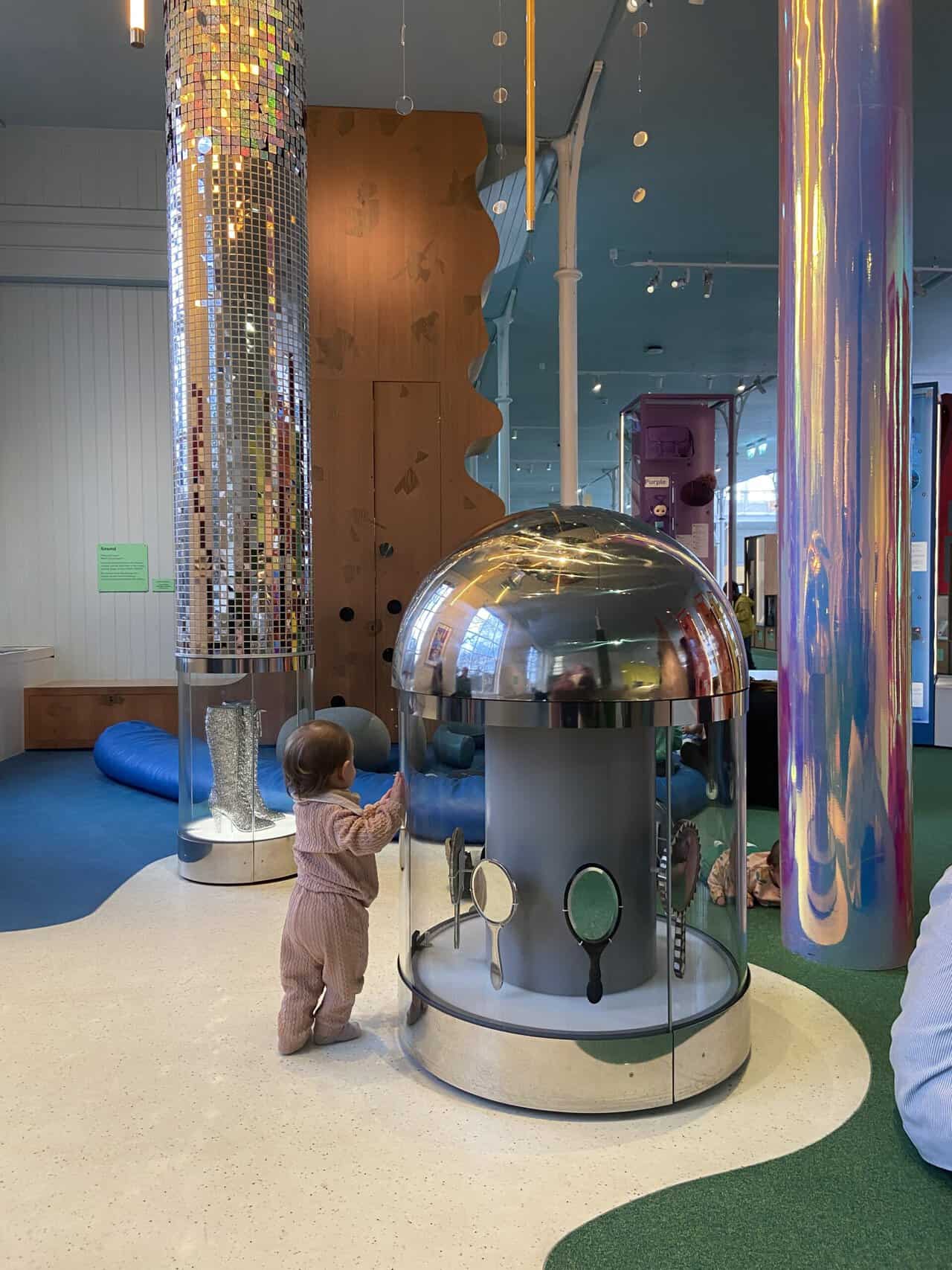 20+ Things to Do in London With a Baby 13 Baby C in the 0-2 year old room at Young V&A children's museum in London. She's standing next to a display of mirrors with shiny poles in front of her. Visiting Young V&A is a free and fun thing to do in London with a baby.