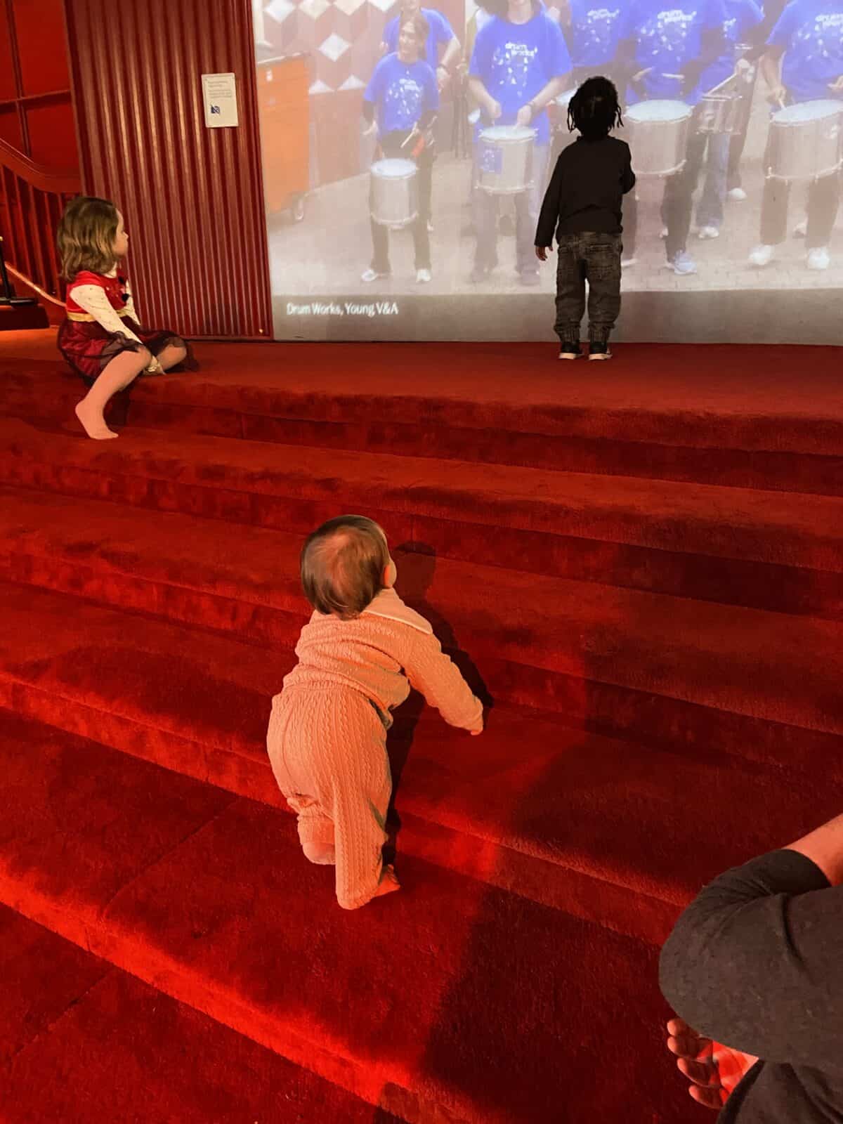 20+ Things to Do in London With a Baby 16 Baby C at the Young V&A children's museum in London. She's in the stage room on the red velvet stairs, looking up at the screen and two other kids.