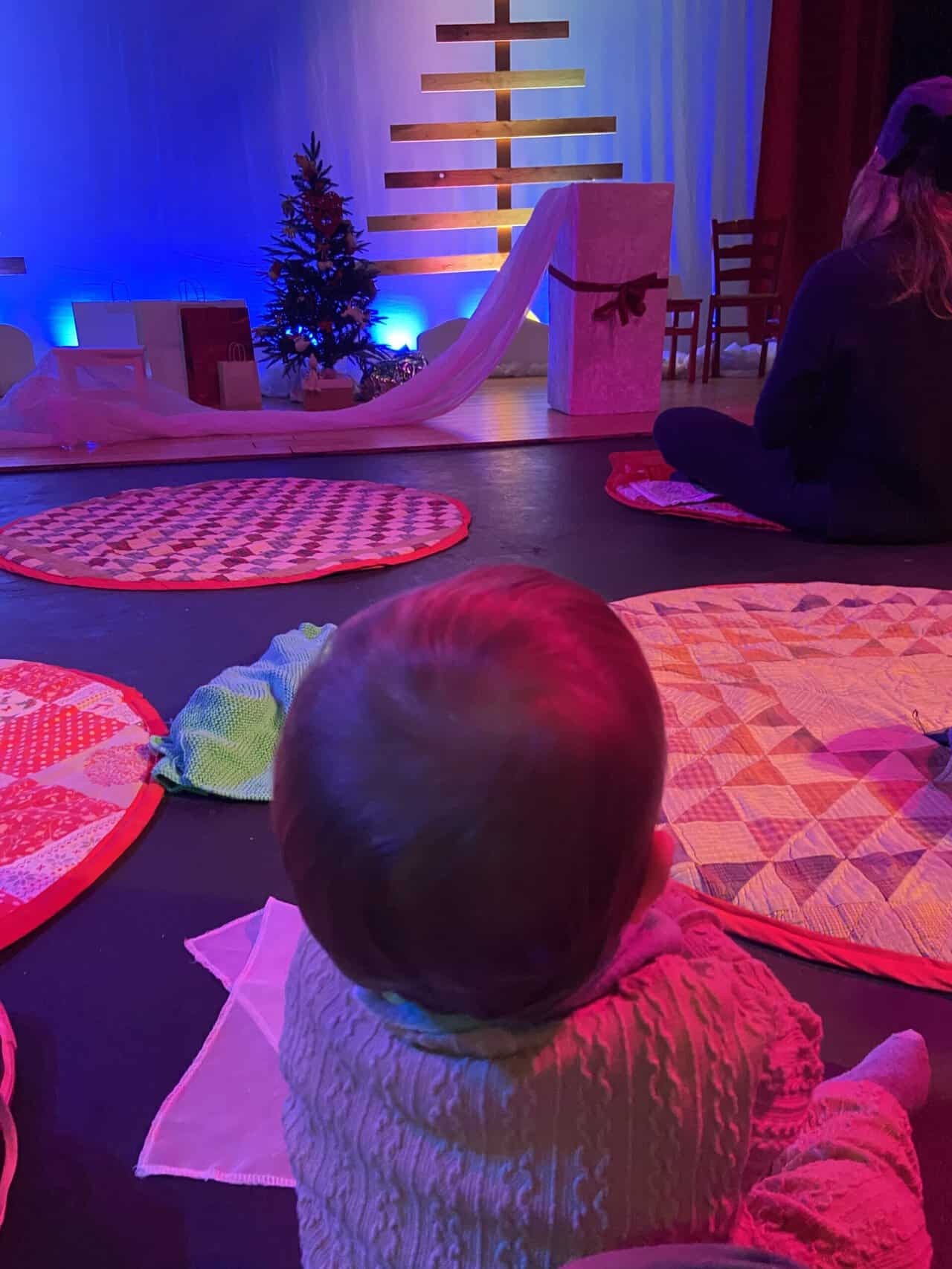 20+ Things to Do in London With a Baby 3 Baby C sitting on the floor of the Unicorn Theatre in London ready to watch Scrunch, a Christmas show for babies. There are cushions on the floor and the stage in front of her has a Christmas tree and presents on it.