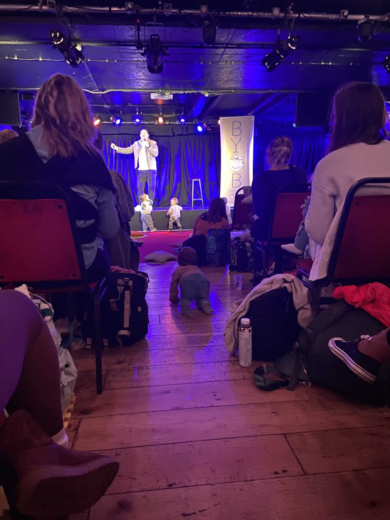 20+ Things to Do in London With a Baby 17 At a BYOB (bring your own baby) comedy show at the Komedia in Brighton, England. A comic stands on stage with a mic in his hands while two babies stand in front of the stage and Baby C crawls down the aisle.