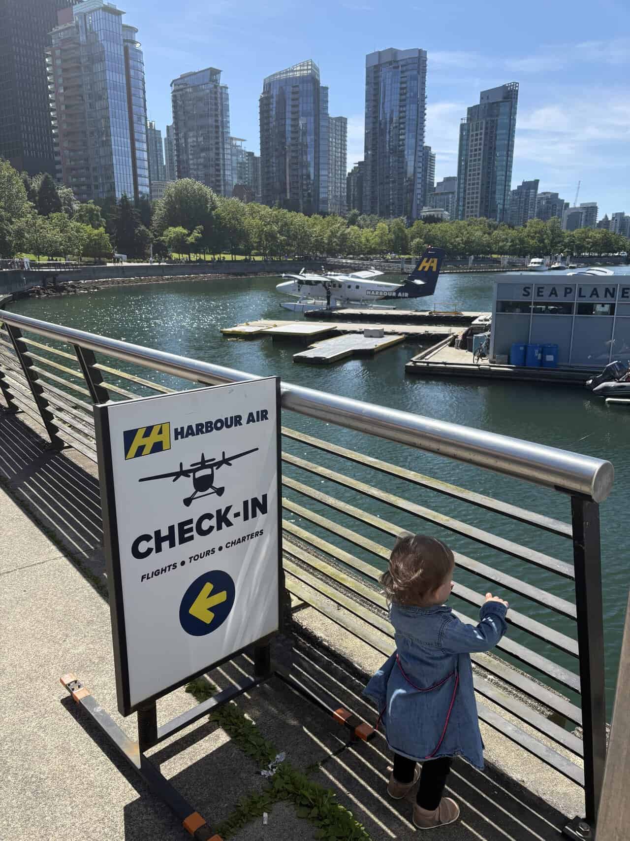 Should You Take a Seaplane to Victoria from Vancouver? (Harbour Air Review) 6 Baby C standing at Vancouver Harbour next to a sign that says "Check In" with seaplanes on the water in the distance