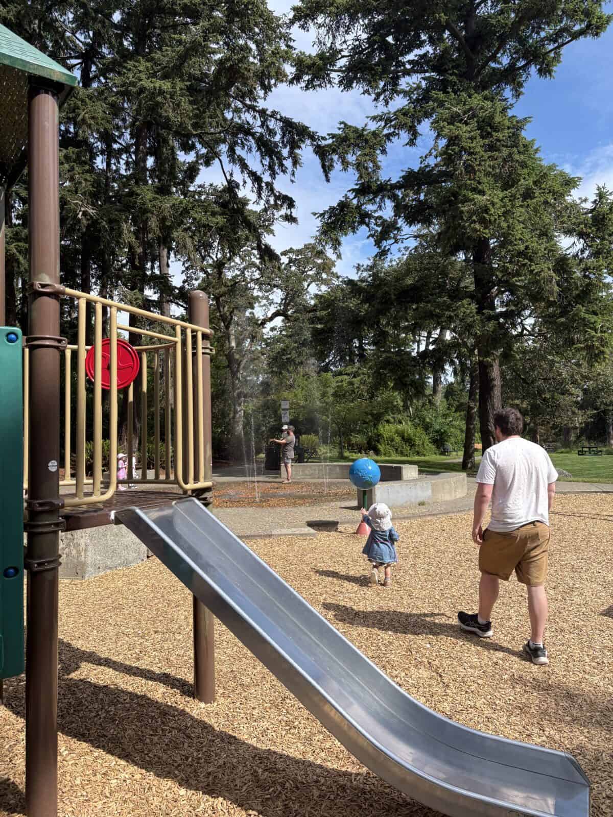 Colin and Baby C running off to the splash pad with a slide in front of them at the Beacon Hill Park North Playground in Victoria, BC