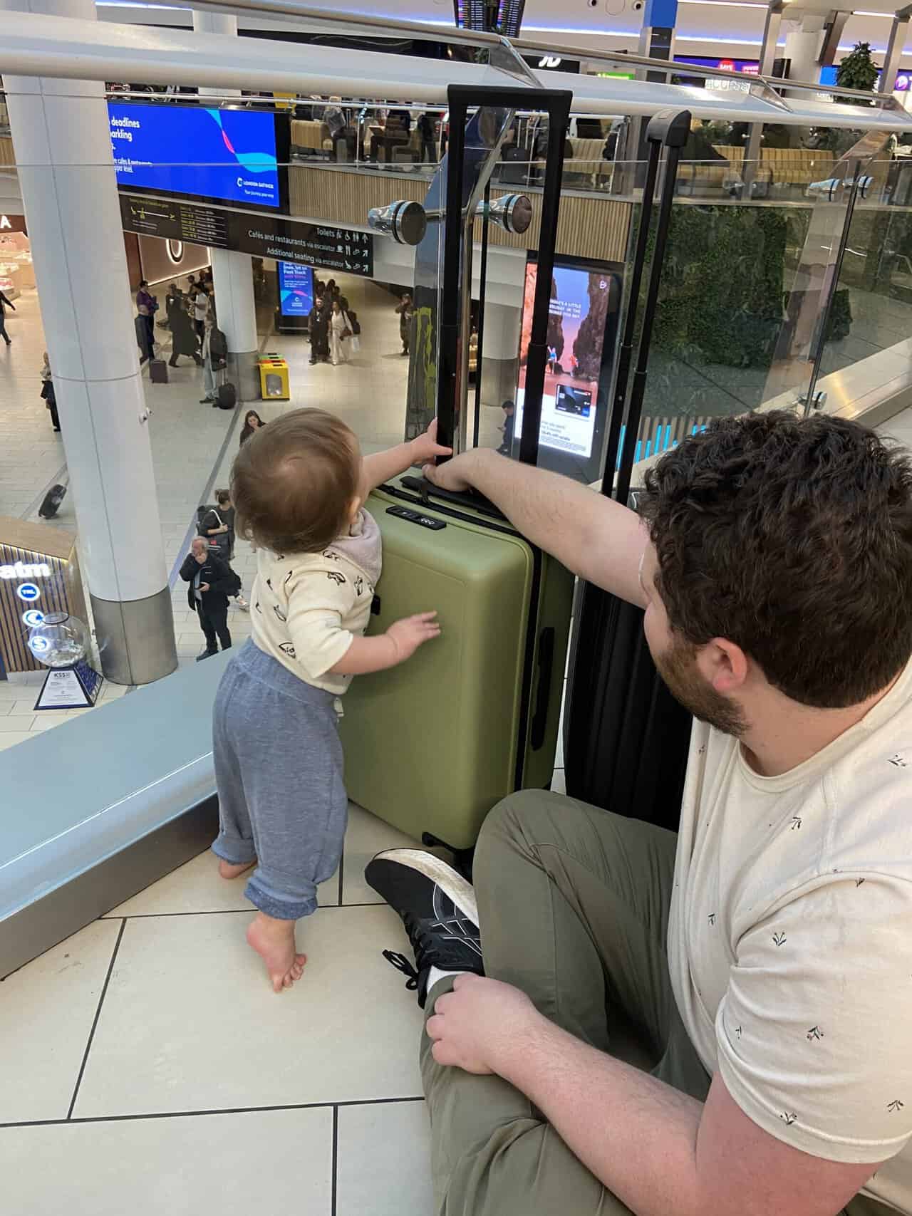 Tips for Flying With a Baby: From Booking to Baggage Claim 23 Baby C pushing our carry-on suitcases at Gatwick Airport while Colin holds onto them