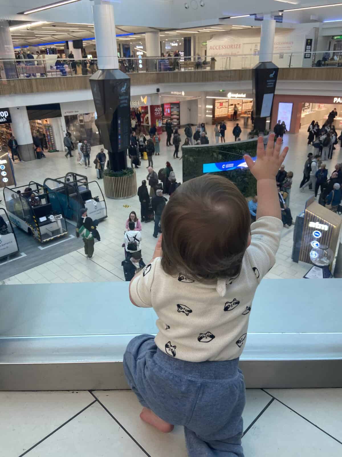 Tips for Flying With a Baby: From Booking to Baggage Claim 33 Baby C sitting on the floor at Gatwick Airport looking through a glass wall at people walking by