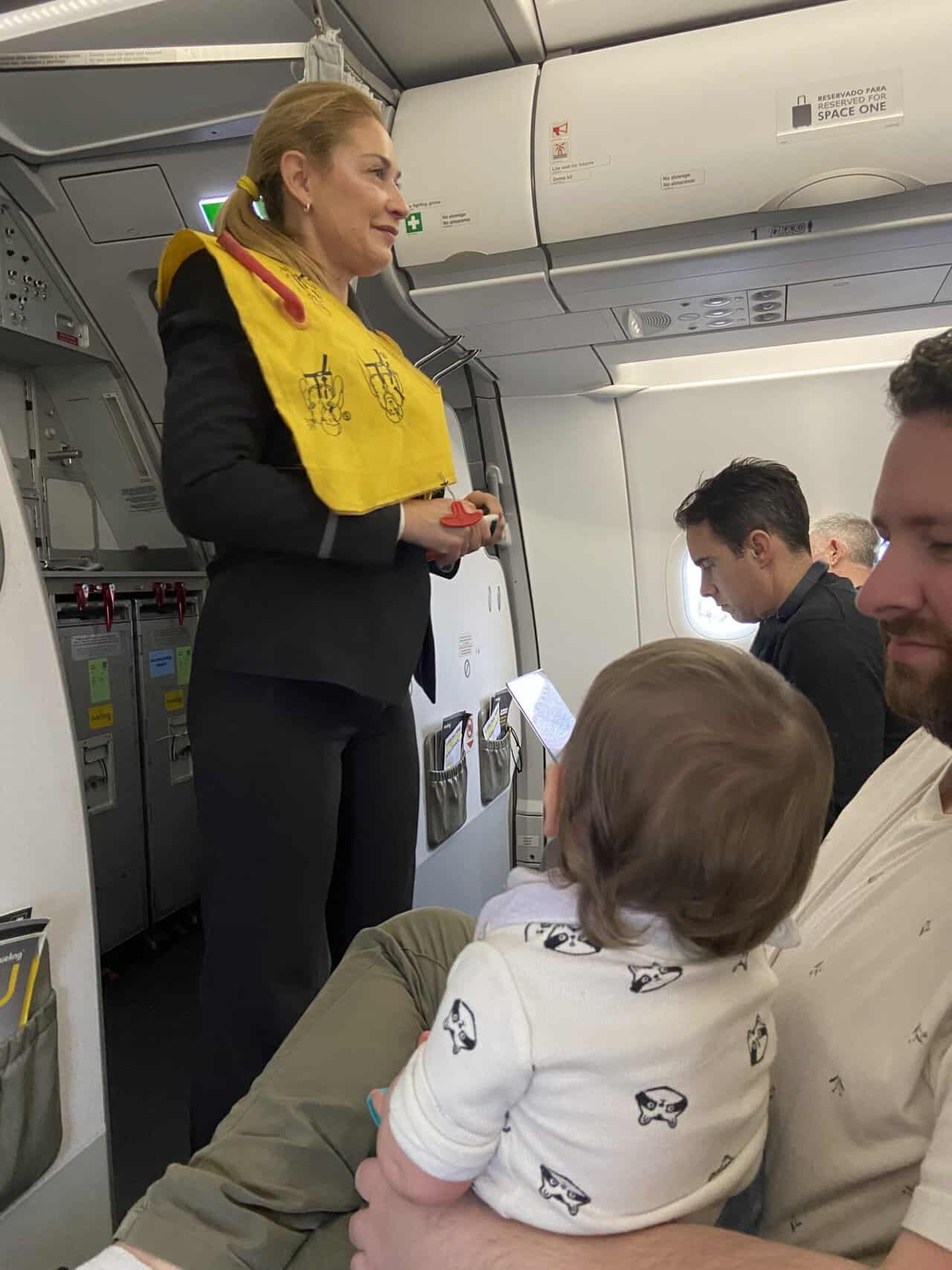 Tips for Flying With a Baby: From Booking to Baggage Claim 37 Baby C sitting on Colin's lap and watching a flight attendant do the safety demo before takeoff