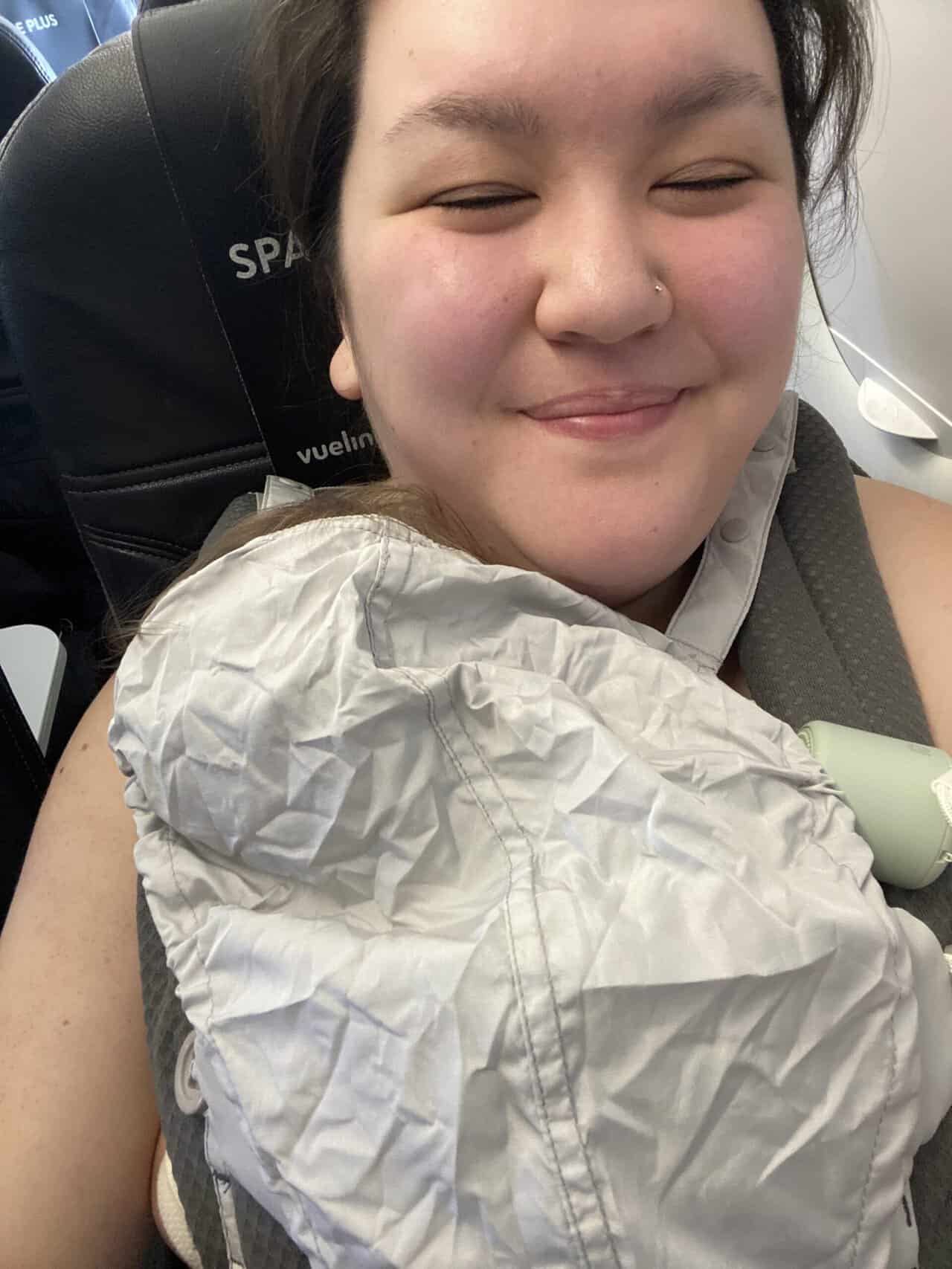 Tips for Flying With a Baby: From Booking to Baggage Claim 16 Riana wearing Baby C in a carrier on the plane with the cover over her head during a nap