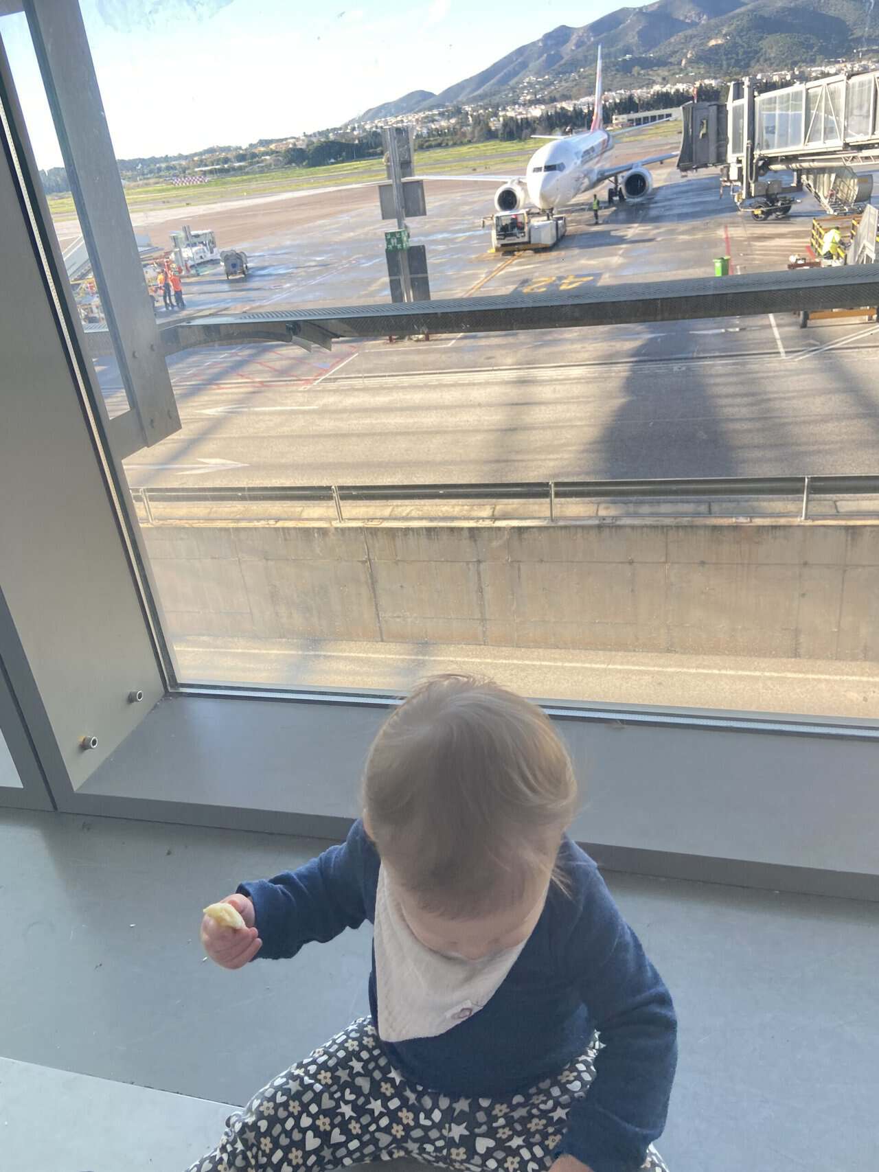 Tips for Flying With a Baby: From Booking to Baggage Claim 27 Baby C waiting at an airport gate, sitting by the window, with a plane behind her