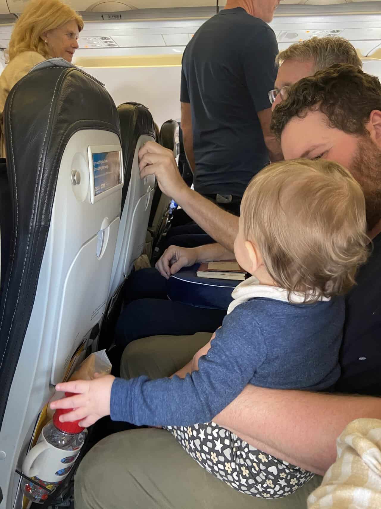 Tips for Flying With a Baby: From Booking to Baggage Claim 36 Baby C sitting on Colin's lap on an airplane while other passengers board