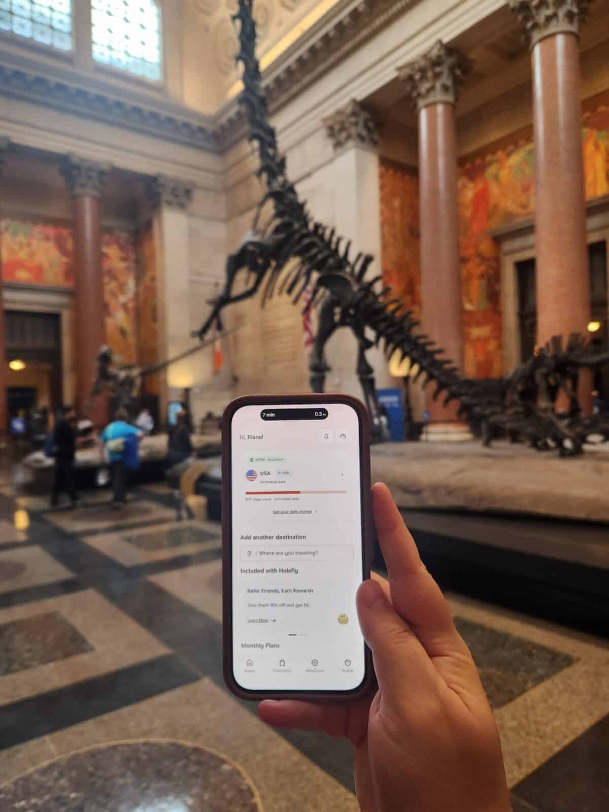 Riana holding her phone showing off the Holafly app with her USA eSIM in front of the big dinosaur at the American Museum of Natural History in NYC